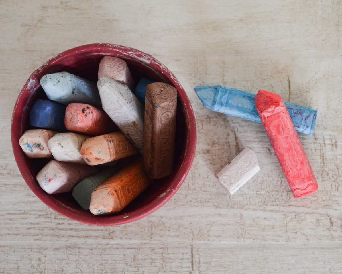 A red bowl filled with various colored chalk sticks sits on a wooden table, capturing the creativity fostered at Excellence In Care family day care. Beside the bowl, three chalk sticks in blue, red, and white lie ready for little hands to express their imagination.