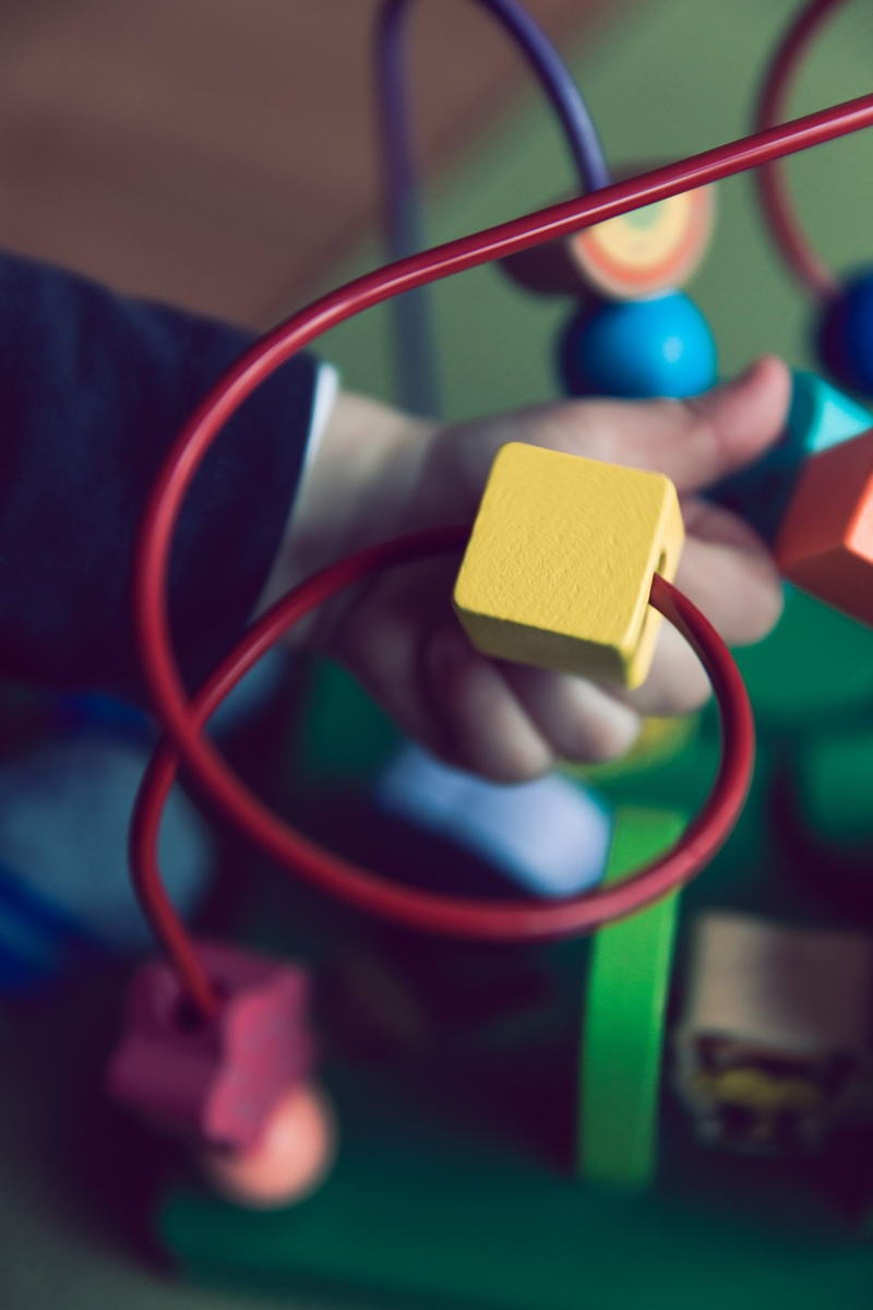 A child's hand is playing with a colorful bead maze toy, guiding a yellow cube along a red wire, embodying the Excellence In Care found at family day care. The toy boasts various shapes and colors designed for sensory and motor skills development.
